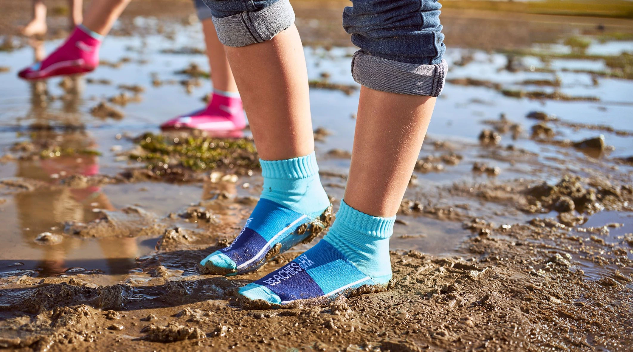 Kinderfüße in blauen Beachies® Socken stehen im Schlick, dahinter ein Kind in pinken Beachies® Socken.