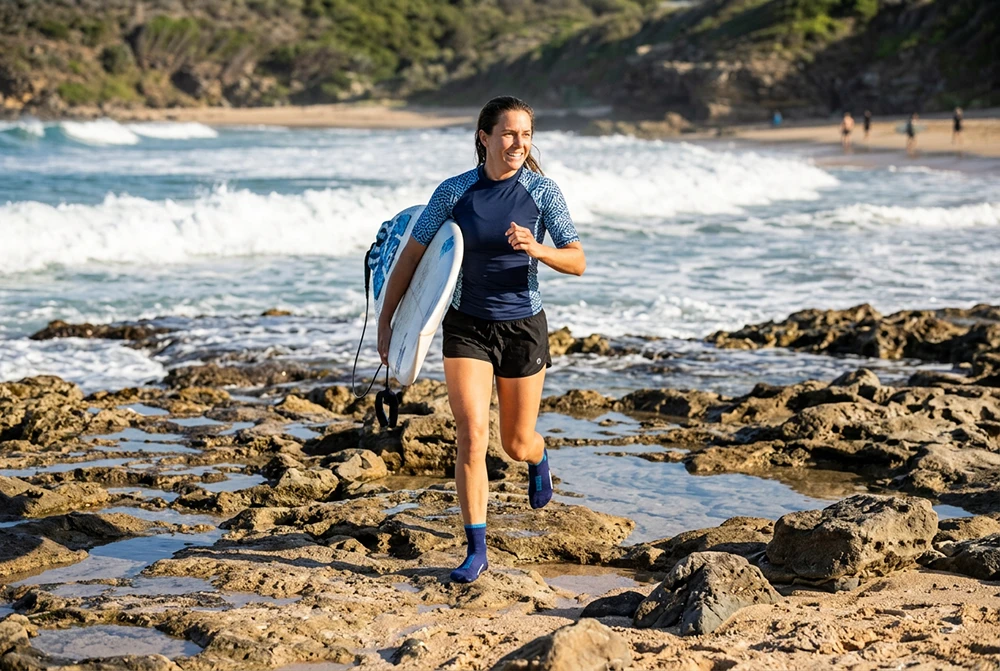 Person läuft mit Surfboard über felsigen Strand und trägt blaue Schwimmsocken.