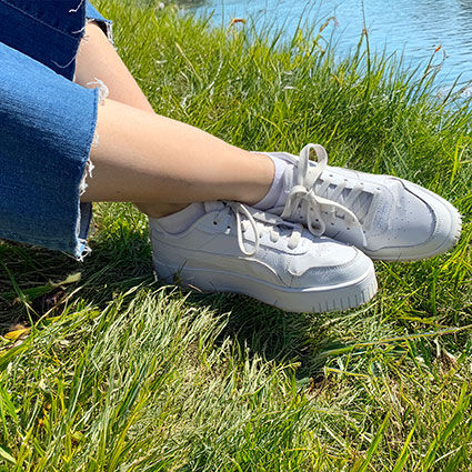 Person in einer Jeans mit Fransen sowie weißen Sneakern mit weißen Sneakersocken auf einer grünen Wiese bei Sonnenschein am Wasser. Beine überkreuzt.