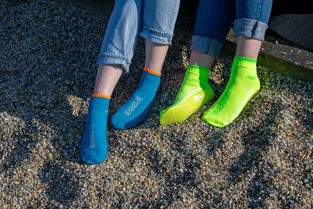 Zwei Personen tragen BEACHIES® Socken in Blau und Neongrün, die Füße auf Kieselsteinen abgestellt.