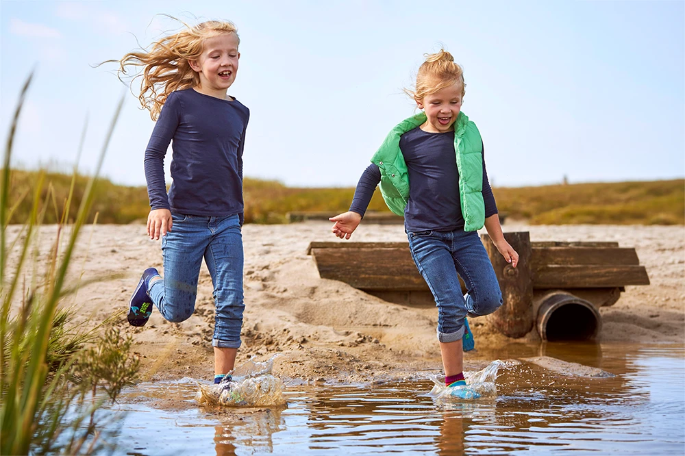 Zwei lachende Kinder in BEACHIES® Socken rennen durch flaches Wasser am Sandstrand.