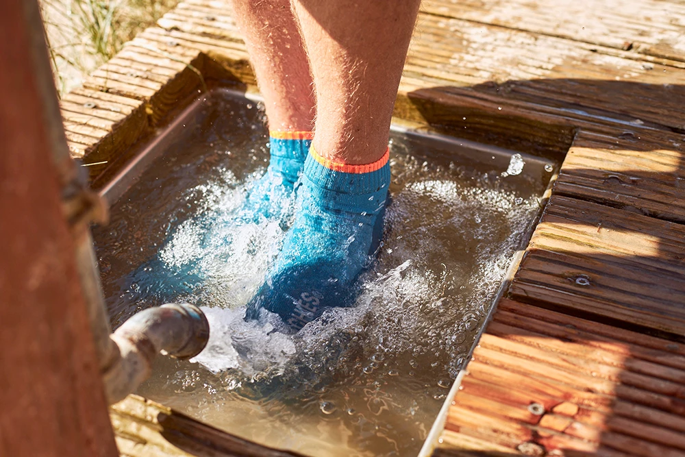 Füße in blauen BEACHIES® Socken werden an einer hölzernen Fußwaschstelle unter spritzendem Wasser abgespült.