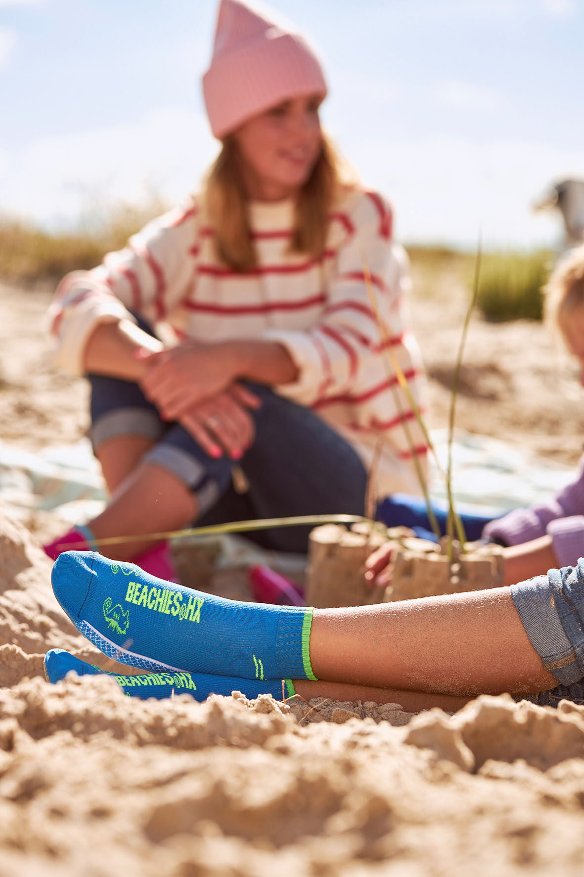 Frau im Sand mit Kindern und Beachies, den Strand- und Wassersportsocken in blau und pink.