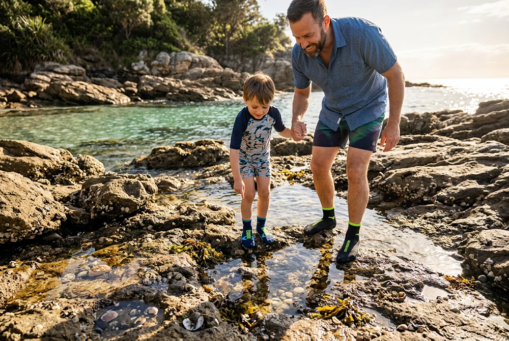 Ein Mann und ein Kind in dunklen BEACHIES® Socken erkunden Hand in Hand einen Gezeitentümpel in einer felsigen Küstenlandschaft