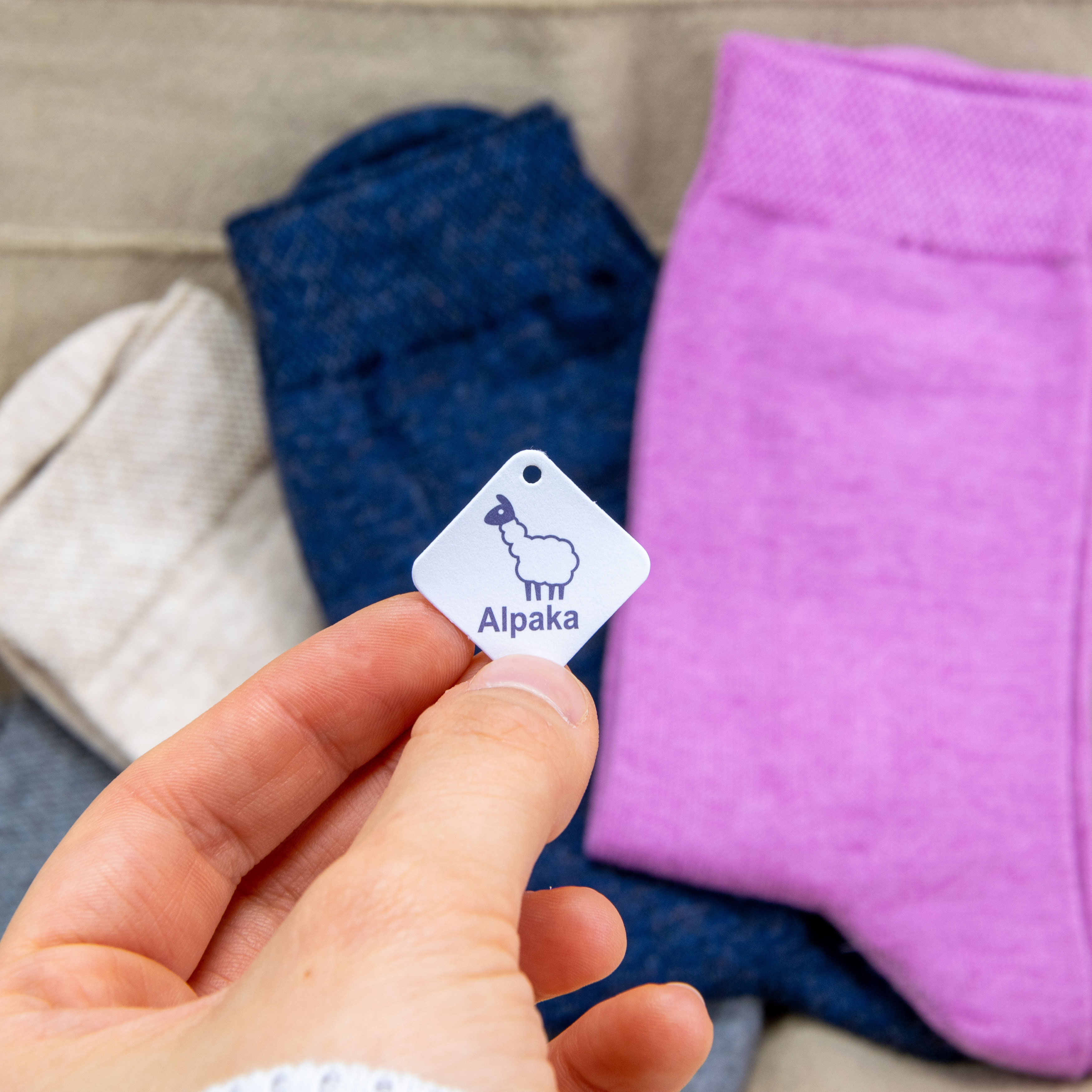 Drei Paar Alpakasocken liegen nebeneinander auf einer Decke. Eine Hand hält ein Alpaka Hang-Tag ins Bild.