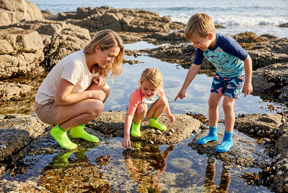Eine Frau und zwei Kinder in neongrünen und blauen BEACHIES® Socken erkunden gemeinsam einen Gezeitentümpel in einer felsigen Küstenlandschaft.