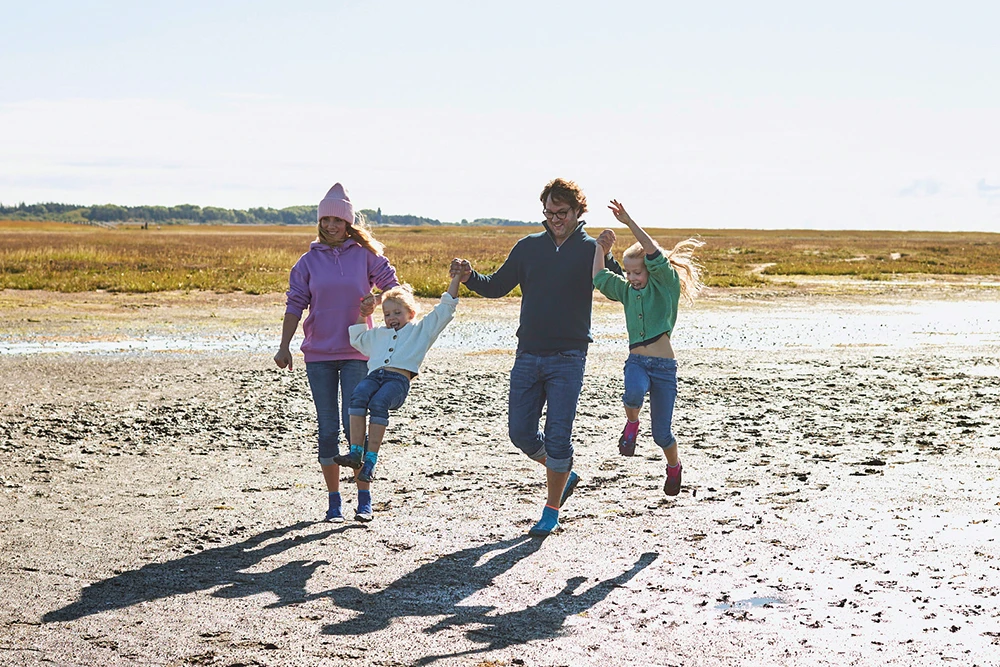 Eine vierköpfige Familie in BEACHIES® Socken läuft lachend Hand in Hand über eine weite Wattlandschaft und schwingt die Kinder in die Luft.