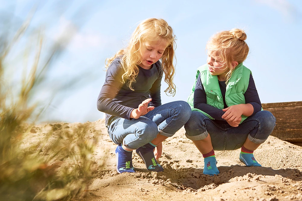 Zwei Kinder in BEACHIES®-Wattsocken hocken im Sand und betrachten etwas am Boden.
