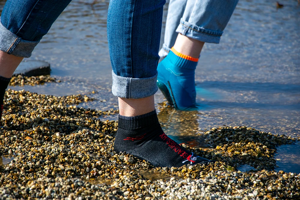 Zwei Personen in aufgekrempelten Jeans tragen BEACHIES® Socken in Schwarz und Blau und stehen auf Kieselsteinen am flachen Wasser.
