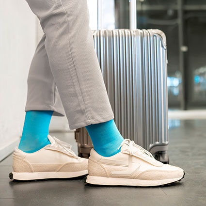 Beine einer Person bekleidet mit grauer Hose und blauen Bionmovesocken in Sneakern neben einem Koffer auf Reisen.