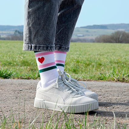 Beine einer Frau bekleidet mit weißen Tennissocken mit Herzmotiv und Ringeln, weißen Sneakern und grauer Jeanshose auf einem Gehweg. Im Hintergrund Natur und Wiese.