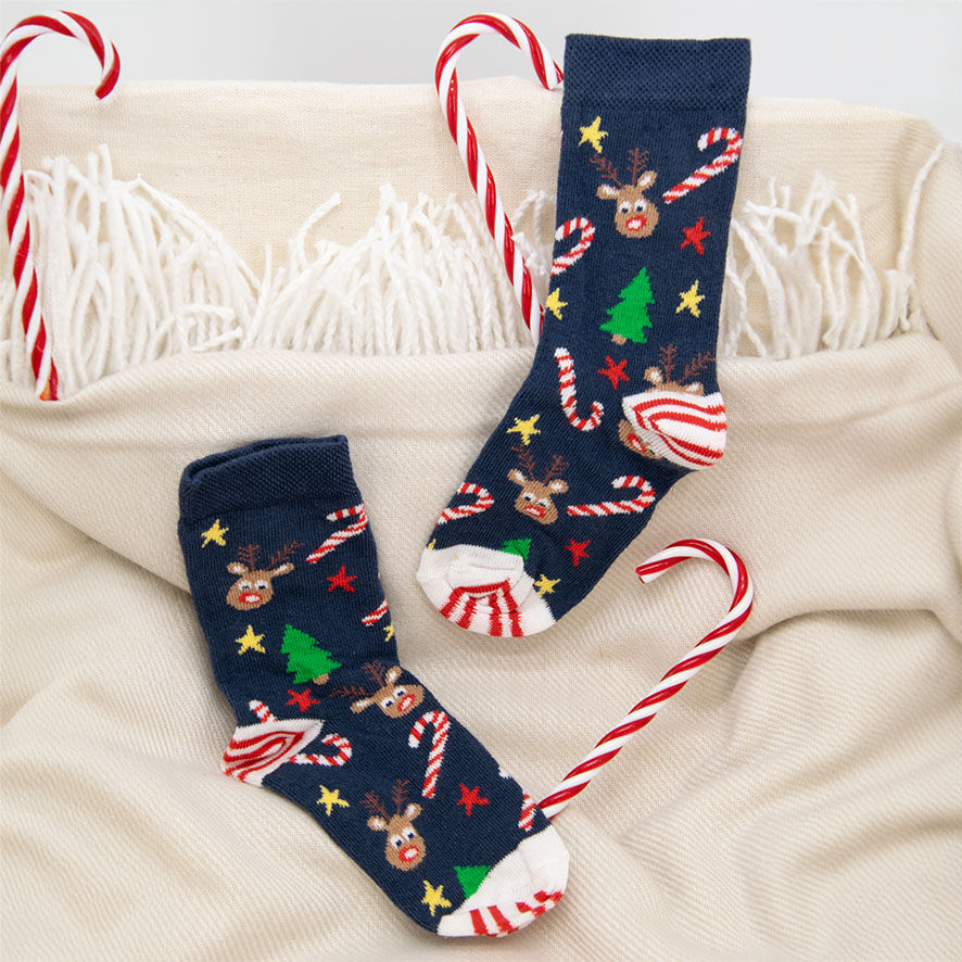 Weihnachtssocken dunkelblau/marine/navy mit Motiv Zuckerstangen, Rentier, Sterne und Weihnachtsbäume in bunten Farben liegen auf einer Decke in weiß/beige/cremefarbig. Daneben liegen Zuckerstangen in rot/weiß.